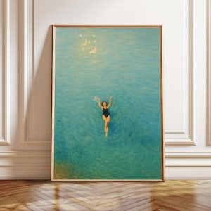 May include: A framed art print depicts a person swimming in turquoise water, arms raised. The person wears a black swimsuit. The artwork is in a gold-colored frame, displayed against a white wall and wooden floor.
