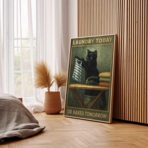 May include: A framed poster features a black cat holding a blue and white striped garment, with an iron and folded laundry on a table. Text reads "LAUNDRY TODAY OR NAKED TOMORROW". The artwork has a vintage, humorous theme.