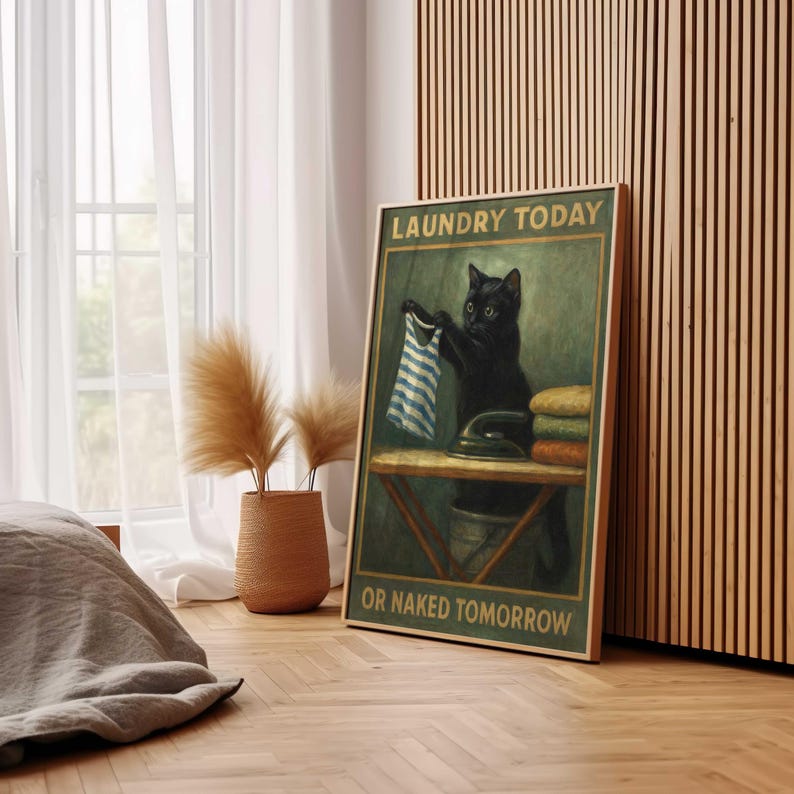May include: A framed poster features a black cat holding a blue and white striped garment, with an iron and folded laundry on a table. Text reads "LAUNDRY TODAY OR NAKED TOMORROW". The artwork has a vintage, humorous theme.
