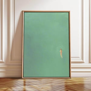 May include: An abstract art print featuring a swimmer in a turquoise body of water. The artwork is framed in a gold-colored frame and is displayed against a white wall and wooden floor.