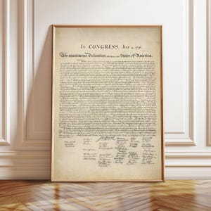 May include: Framed print of the Declaration of Independence. The document is displayed on aged parchment paper with black ink. The text includes the date July 4, 1776, and signatures at the bottom. The frame is a light brown color.