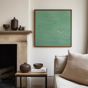 以下が含まれることがあります： 緑色の水中で泳ぐ2人の人物を描いた額入りのアートワークがあるリビングルームのシーン。木製のサイドテーブルには、濃い茶色の花瓶とボウルが置かれています。さまざまな茶色のセラミック容器が石の暖炉にあり、テクスチャード加工のクッションが付いたベージュのソファが前景にあります。