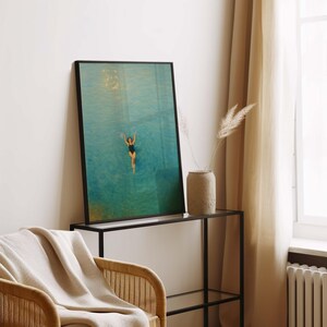 May include: Framed art print featuring a person swimming in turquoise water. The artwork is displayed in a black frame and is set against a neutral-toned wall. A beige throw blanket rests on a wicker chair.