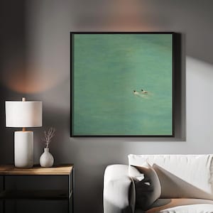 以下が含まれることがあります： ターコイズブルーの海で泳ぐ2人の姿を描いた額入りのアートプリント。アートワークは、白いソファの上にある灰色の壁に飾られています。白いランプと小さな花瓶が木製のサイドテーブルに置かれています。