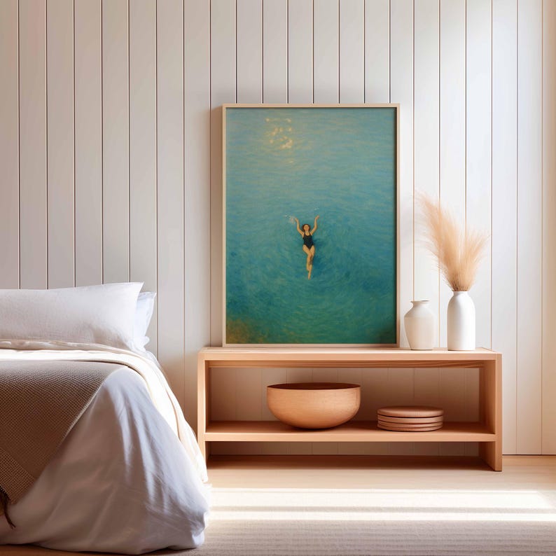 May include: A framed art print depicts a person swimming in turquoise water. The artwork is displayed above a wooden console table with decorative vases, a wooden bowl, and plates. The room features a bed with white linens and a neutral color palette.