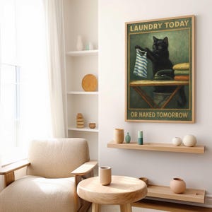 May include: A framed print featuring a black cat holding a striped shirt above an ironing board. The text reads "LAUNDRY TODAY OR NAKED TOMORROW". The artwork has a vintage aesthetic, with a green and brown color palette. The print is displayed in a living room setting.