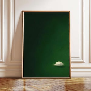 May include: A framed art print featuring a deep green background with a single, fluffy white cloud. The artwork is displayed in a gold-colored frame and leans against a white wall and wooden floor.