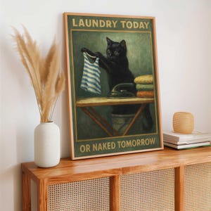 May include: A framed print featuring a black cat holding a striped shirt over an ironing board. The print reads "LAUNDRY TODAY OR NAKED TOMORROW" in a vintage style. The cat is next to a stack of folded towels and an iron.