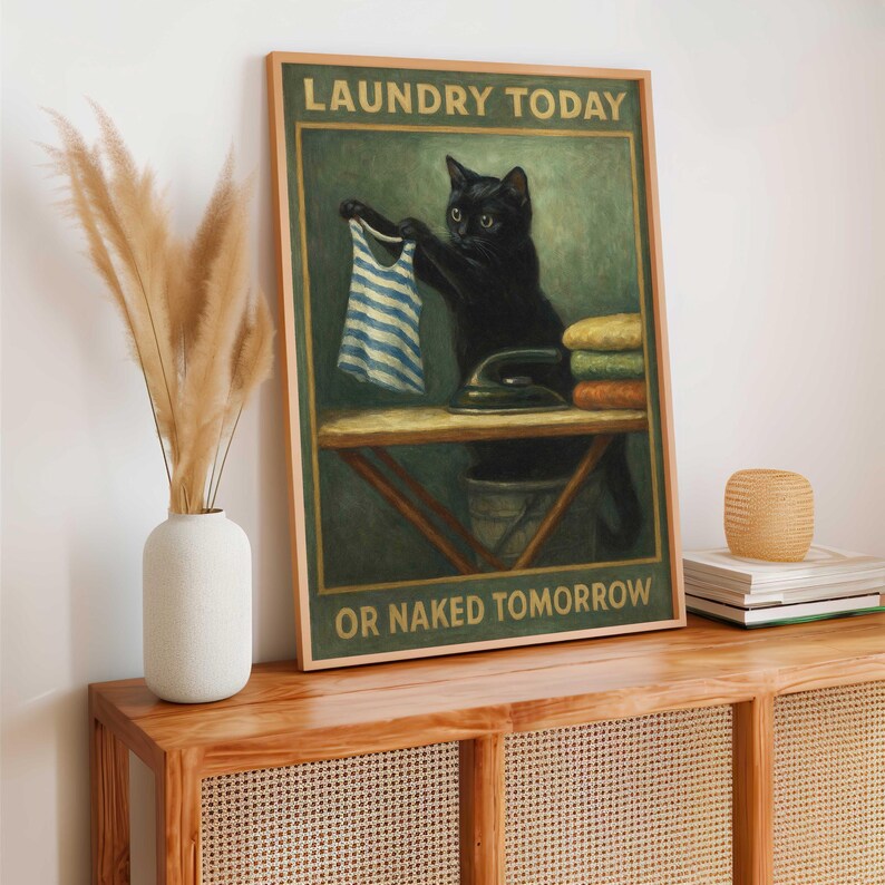 May include: A framed print featuring a black cat holding a striped shirt over an ironing board. The print reads "LAUNDRY TODAY OR NAKED TOMORROW" in a vintage style. The cat is next to a stack of folded towels and an iron.