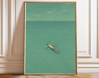 May include: A framed art print depicts a person floating in turquoise water under a pale green sky. The person is wearing a white swimsuit. The artwork is in a gold frame and leans against a white wall and wooden floor.