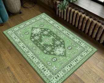 May include: A rectangular green rug with an intricate geometric pattern. The rug features a central dark green design surrounded by lighter green and white borders. The rug is placed on a wooden floor.