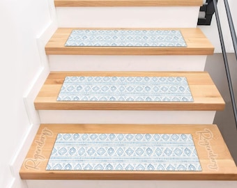 Op de afbeelding: Houten trap met drie treden, elk voorzien van een blauw en wit patroon trapmat. De treden hebben een geometrisch ontwerp. De stootborden zijn wit, wat zorgt voor een strakke, moderne uitstraling.