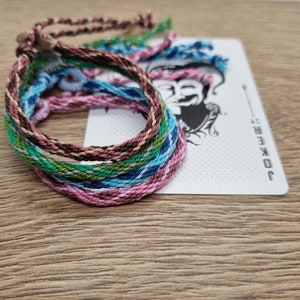 Può includere: Braccialetti dell'amicizia intrecciati in vari colori, tra cui rosa, verde, blu e marrone. I braccialetti sono impilati su una carta da gioco con un disegno di jolly.