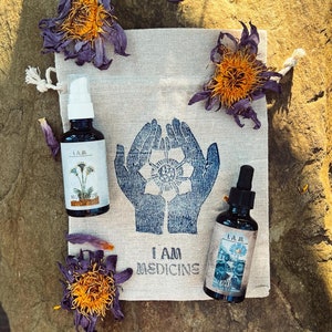 Könnte beinhalten: Zwei kleine Glasflaschen mit Etiketten und blauen und weißen Illustrationen, die auf einem weißen Stoffbeutel mit dem Aufdruck "I AM MEDICINE" liegen. Der Beutel liegt auf einem grauen Stein mit getrockneten lila Blumen.