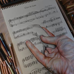 May include: A realistic colored pencil drawing on paper featuring a hand appearing to lift a section of sheet music titled Clair de Lune by Claude Debussy. The artwork displays intricate musical notation and a detailed hand with skin tones.