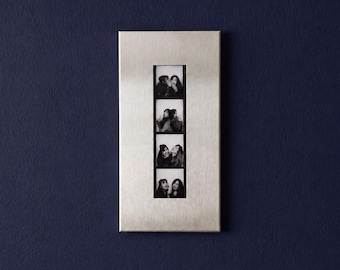 Puede incluir: Un marco de fotos rectangular de metal plateado cepillado con un recorte vertical que muestra una tira de cuatro fotografías en blanco y negro. Cada marco individual en la tira captura a dos personas posando juntas con diversas expresiones.
