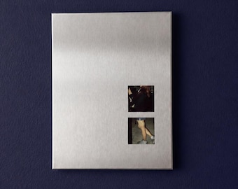 Puede incluir: Una placa de pared rectangular de metal plateado cepillado con dos pequeños recortes cuadrados dispuestos verticalmente. El recorte superior muestra una imagen oscura, mientras que el inferior revela una vista parcial de piernas y pies con calzado claro.