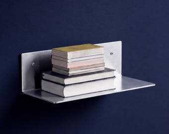 May include: A brushed metal wall-mounted shelf with a flat horizontal surface and a vertical back panel secured by two visible screws. A stack of five books with varying spine colors and thicknesses rests on the shelf surface.
