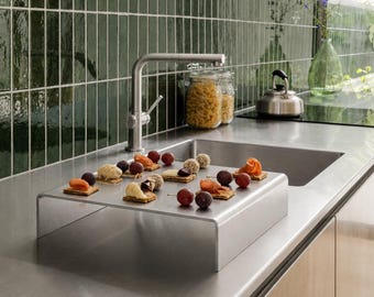 May include: A brushed stainless steel rectangular riser stand sits on a matching metal countertop. The stand holds an assortment of small appetizers, including crackers topped with salmon, cheese, and fresh grapes arranged on wooden skewers.