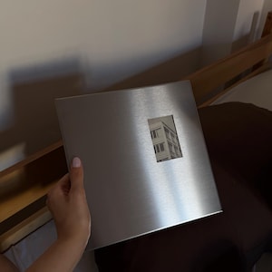 Hand holding a square brushed metallic silver photo album with a small rectangular window showing a picture.