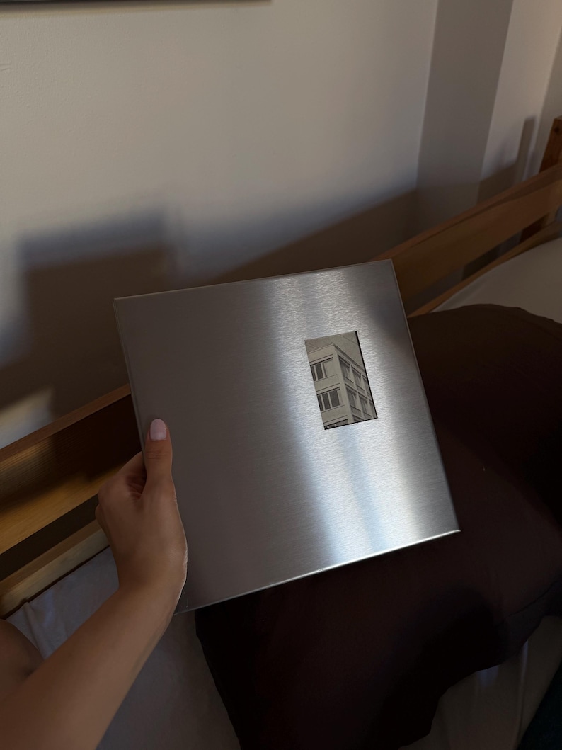 Hand holding a square brushed metallic silver photo album with a small rectangular window showing a picture.