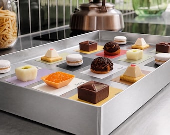 May include: A rectangular brushed metal tray containing an assortment of small desserts, including chocolate squares, macarons, cream puffs, and pyramid-shaped cakes, each resting on a small, colorful square card with printed text labels.