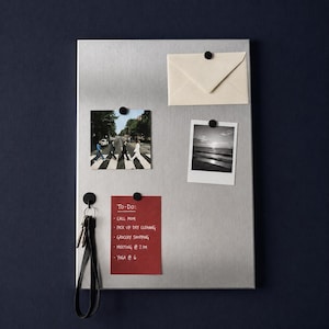 May include: A rectangular brushed silver metal magnetic board featuring a cream envelope, a color photograph of people crossing a street, a black and white sunset photograph, and a red to-do list with white text, all held by small black magnets.