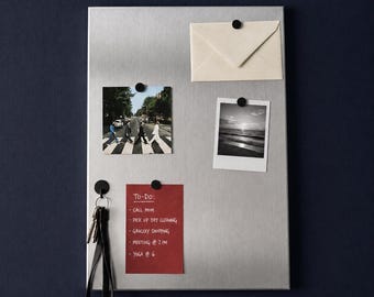 Puede incluir: Un tablero magnético rectangular de metal plateado cepillado con un sobre color crema, una fotografía en color de personas cruzando una calle, una foto en blanco y negro de un atardecer y una lista de tareas roja con texto blanco, sujetos por pequeños imanes negros.
