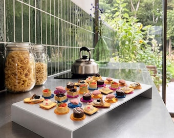 May include: A rectangular white acrylic riser stand displays an assortment of small, colorful bite-sized desserts on individual circular bases. The treats feature various toppings including berries, cream, and chocolate, arranged on a kitchen counter.