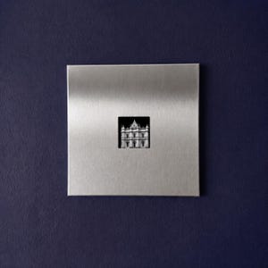 May include: A square, brushed silver metal wall plaque featuring a centered, small, black and white photographic cutout of a classical building with a prominent arched entrance and multiple levels, set against a dark, solid navy blue background.