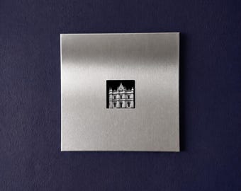 Puede incluir: Una placa de pared cuadrada de metal plateado cepillado que presenta un pequeño recorte fotográfico en blanco y negro centrado de un edificio clásico con una entrada arqueada prominente y varios niveles, sobre un fondo azul marino.