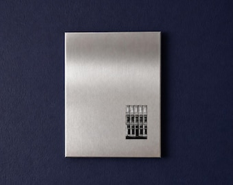 May include: A rectangular brushed metal plaque with a vertical orientation. The surface features a fine, linear grain texture. A small, black and white photograph of a multi-story building facade with multiple windows is centered near the bottom.