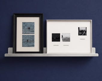 May include: A silver metal floating shelf holding two picture frames. The left frame is black with two stacked photos of a person swimming. The right frame is white with three small black and white photos and text captions underneath each image.