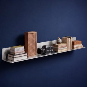 May include: A long, horizontal brushed metal wall shelf holding a stack of books, a tall wooden box with a grid pattern, a black vintage camera, a small ceramic vase, and several additional books arranged in a neat, organized display.