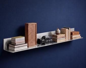 Puede incluir: Una estantería de pared larga y horizontal de metal cepillado que sostiene una pila de libros, una caja de madera alta con patrón de cuadrícula, una cámara vintage negra, un pequeño jarrón de cerámica y varios libros adicionales.