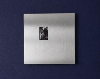 Puede incluir: Una placa de pared cuadrada de metal plateado cepillado con una pequeña fotografía rectangular en blanco y negro de dos personas abrazándose centrada. La superficie metálica tiene una textura de grano horizontal y un acabado liso.