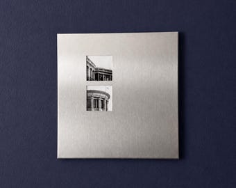 May include: A square, brushed silver metal wall plaque featuring two small, vertically stacked square cutouts. Each cutout reveals a black and white photograph of architectural building details, including columns and a curved structure.