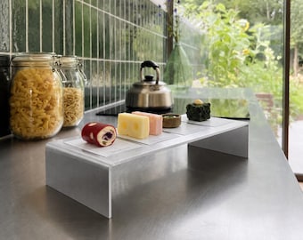 May include: A rectangular brushed metal display stand holds four small food items on individual white cards. The items include a red cylindrical roll, a yellow block of Swiss cheese, a pale pink square, and a small dark green textured square.