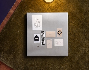 May include: A square brushed metal magnetic board featuring various items attached with magnets, including a drawing of a bottle and glass, a black envelope, a photo strip, a small beige envelope, a patterned card, and a note reading you are always welcome home.