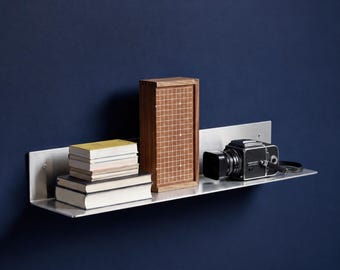 May include: A brushed metal wall-mounted shelf holding a stack of books with light-colored covers, a tall rectangular wooden box featuring a grid pattern on its front face, and a black vintage-style camera with a lens hood attached to the side.