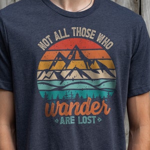 May include: A dark blue t-shirt with a vintage-style graphic of mountains and a sunset. The text "Not all those who wander are lost" is printed on the shirt.