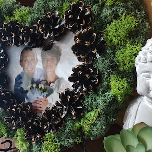 Op de afbeelding: Een hartvormige fotolijst met een foto van een stel, omringd door groen mos en zwarte dennenappels met gouden accenten. Een kleine Boeddha-beeld en een vetplant staan op de achtergrond.
