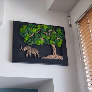 Può includere: Un'opera d'arte da parete incorniciata con un rilievo 3D di un elefante e un albero. L'elefante è color bronzo e l'albero ha una chioma verde muschiosa. L'opera è ambientata su uno sfondo nero.