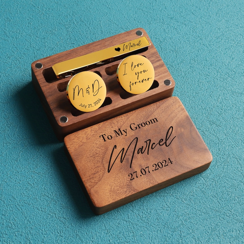 May include: A wooden box with a gold tie bar and two gold cufflinks. The cufflinks have the text "M&D July 27, 2024" and "I love you forever". The box has the text "To My Groom Marcel 27.07.2024".