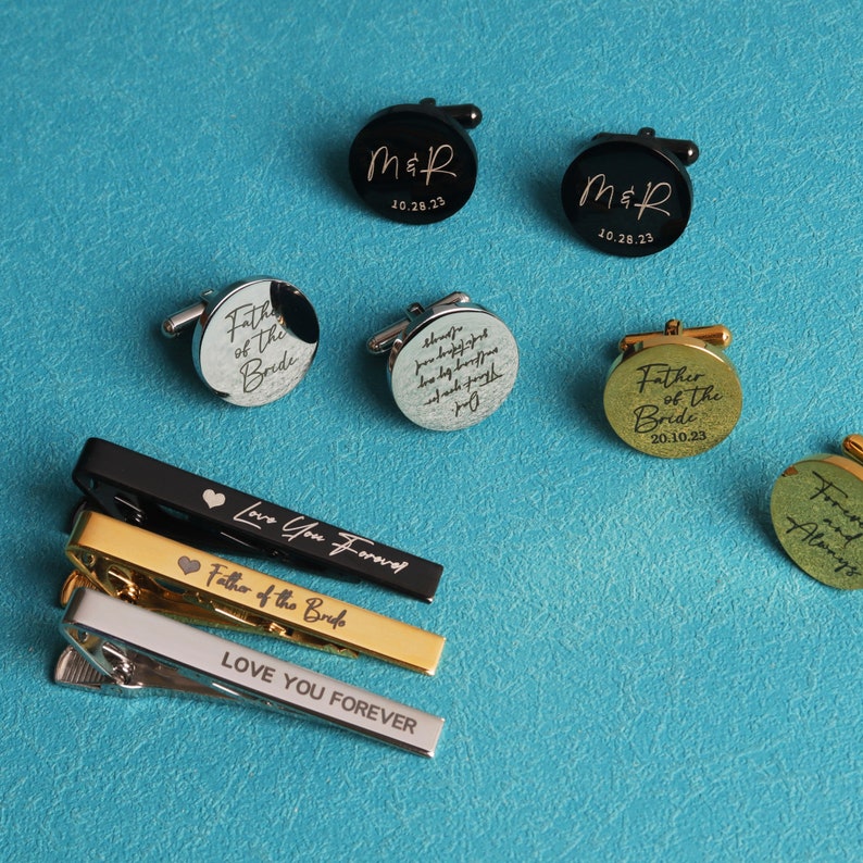 May include: A set of cufflinks and tie clips with engraved messages. The cufflinks are black, silver, and gold, and the tie clips are black, gold, and silver. The messages on the cufflinks include "Father of the Bride", "M&R 10.28.23", and "Always", while the tie clips have the messages "Love You Forever", "Father of the Bride", and "Love You Forever".