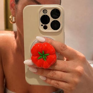May include: A cream-colored phone case with a red tomato-shaped phone grip. The tomato grip has green leaves and is held by a person. The phone case has a camera bump with three black lenses.