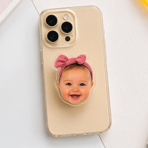 May include: A clear phone case with a smiling baby face pop-socket. The pop-socket features a baby with a pink bow headband. The phone is gold in color.