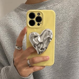 Op de afbeelding: Een gele telefoonhoes met een zilveren hartvormige grip. De hoes heeft een zwart cameramodule en wordt vastgehouden door een persoon met zilveren ringen. De telefoonhoes heeft een effen gele kleur.