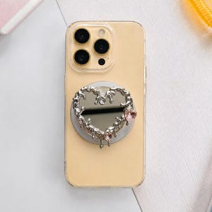 May include: A clear phone case with a silver heart-shaped phone grip. The grip has a mirrored center, embellished with clear and pink rhinestones. The phone is a light yellow color.