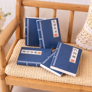 Puede incluir: Pequeños libros azules en miniatura con detalles blancos y texto en idioma extranjero, dispuestos sobre una pequeña silla de madera. Los libros tienen una encuadernación tradicional y probablemente sean para una casa de muñecas o una exhibición similar.
